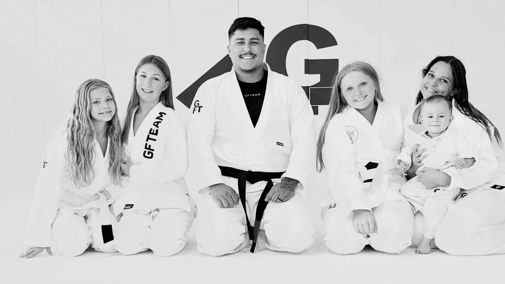 A GFTEAM APEX black belt instructor kneeling on the mat with four young students in gis and a baby, in front of the GFTEAM wall logo.